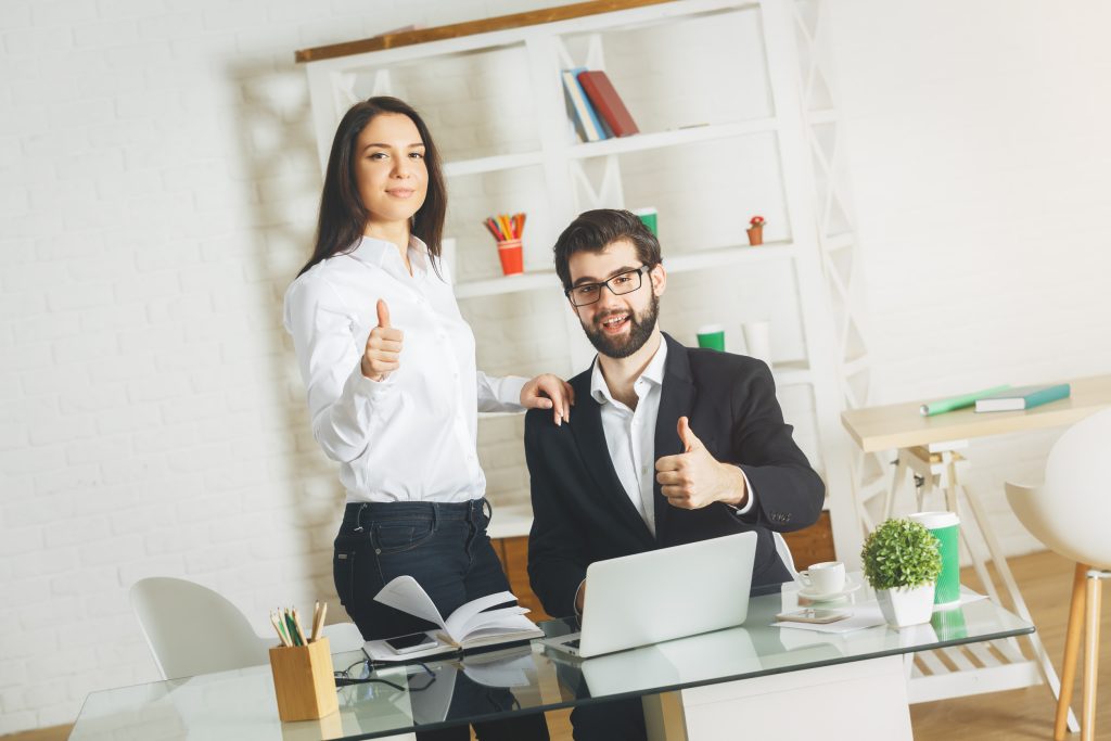 Portrait of attractive businessman and woman working and showing thumbs up at office desk. Meeting, teamwork, discussion, success and togetherness concept